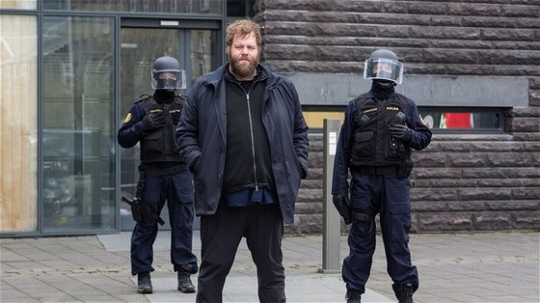 series de advogados - Imagem mostra um homem de preto à frente de dois policiais armados. Todos estão sérios e olham para a câmera